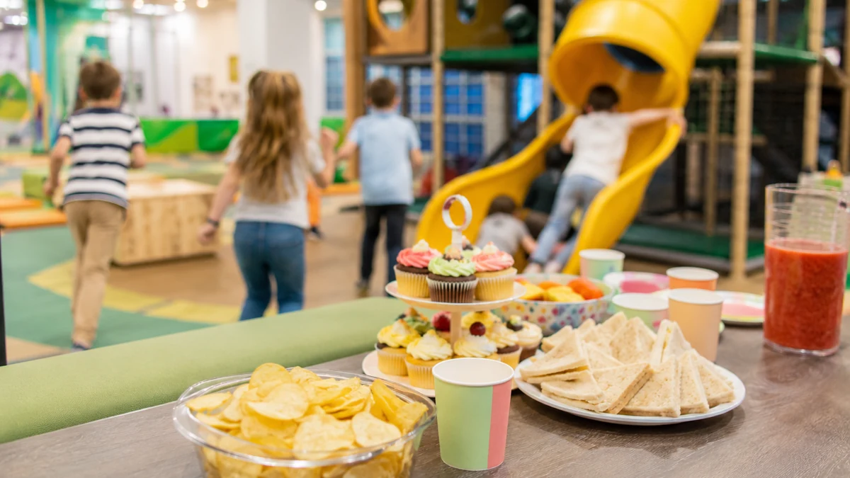 Niños jugando de espaldas en una zona interior de ocio infantil con merienda al fondo